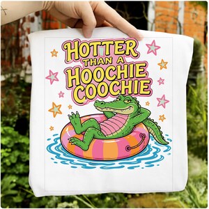 May include: White tote bag with the phrase "Hotter Than a Hoochie Coochie" in yellow and pink lettering. A cartoon alligator relaxes on a pink and orange inner tube in blue water, surrounded by stars.