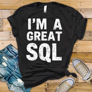 May include: Black t-shirt with the white text "I'M A GREAT SQL". The shirt is displayed with blue jeans and tan trainers on a wooden surface. The t-shirt is knotted at the bottom.