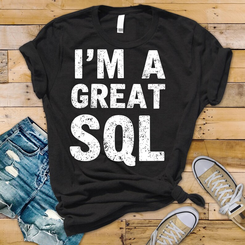 May include: Black t-shirt with the white text "I'M A GREAT SQL". The shirt is displayed with blue jeans and tan trainers on a wooden surface. The t-shirt is knotted at the bottom.