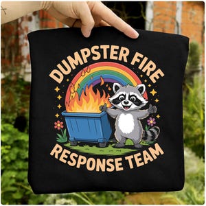 Puede incluir: Bolso tote negro con un diseño de mapache y el texto "Dumpster Fire Response Team". El diseño incluye un contenedor de basura azul en llamas, un arcoíris y un mapache sonriente.
