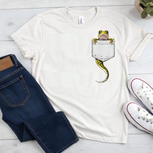May include: Off-white t-shirt with a pocket design featuring a gecko. The gecko is yellow and black, with its head and front legs visible above the pocket. The gecko's tail extends down from the pocket. Dark blue jeans and white trainers are also in the image.