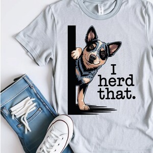 May include: Light gray t-shirt with a graphic of a blue heeler dog peeking out from behind a black line, with the text "I herd that." The shirt is paired with blue jeans and white sneakers.