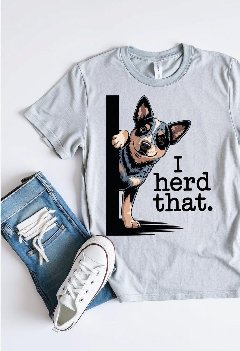 May include: Light gray t-shirt with a graphic of a blue heeler dog peeking out from behind a black line, with the text "I herd that." The shirt is paired with blue jeans and white sneakers.