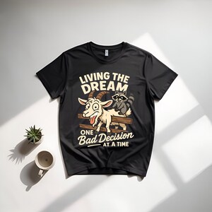 May include: Black t-shirt with a humorous graphic featuring a cartoon goat and raccoon on a wooden fence. The text reads "LIVING THE DREAM" above and "ONE BAD DECISION AT A TIME" below, in a playful font.