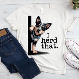 May include: Cream-colored t-shirt featuring a graphic of an Australian Cattle Dog peeking from behind a black line. The text "I herd that." is printed in black. The shirt is paired with blue jeans and white sneakers.