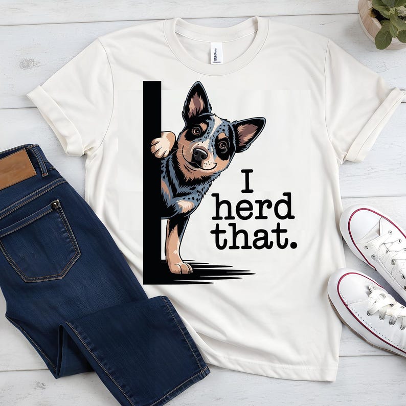 May include: Cream-colored t-shirt featuring a graphic of an Australian Cattle Dog peeking from behind a black line. The text "I herd that." is printed in black. The shirt is paired with blue jeans and white sneakers.