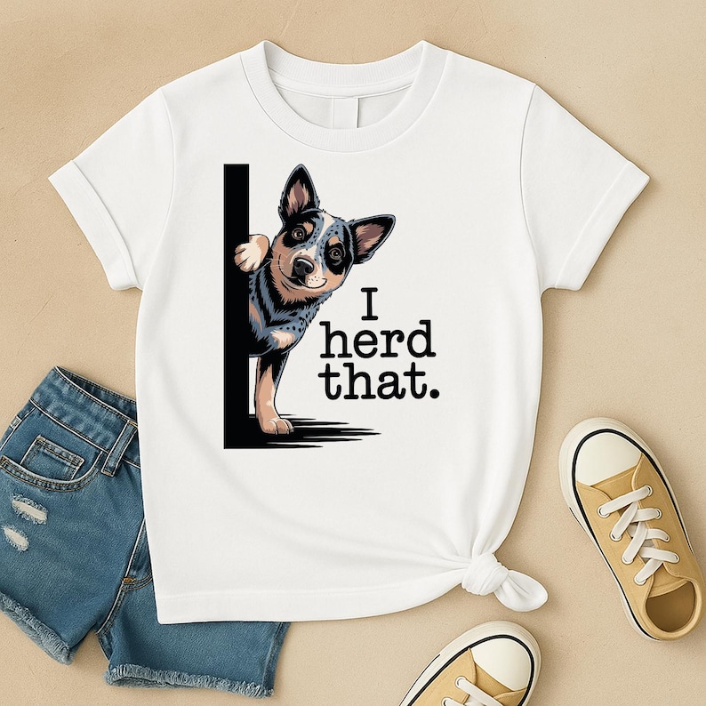 May include: White t-shirt featuring a blue heeler dog graphic peeking from behind a black vertical line. The text "I herd that." is printed in black. The shirt is paired with blue denim shorts and tan sneakers.