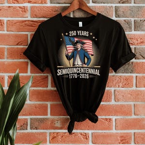 K&ouml;nnte beinhalten: Schwarzes T-Shirt mit einer Grafik einer historischen Figur, die eine amerikanische Flagge h&auml;lt. Das Design enth&auml;lt den Text "250 Years" und "Semiquincentennial 1776-2026". Das T-Shirt h&auml;ngt an einem Holzb&uuml;gel vor einer Backsteinmauer.