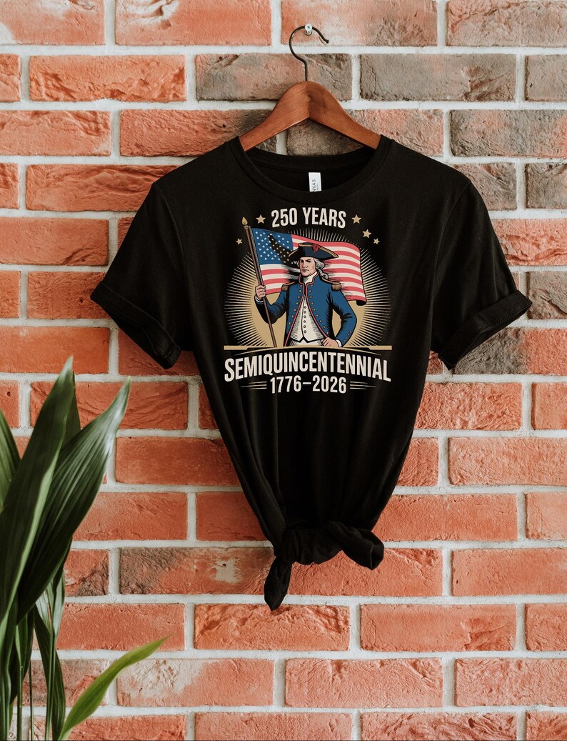 K&ouml;nnte beinhalten: Schwarzes T-Shirt mit einer Grafik einer historischen Figur, die eine amerikanische Flagge h&auml;lt. Das Design enth&auml;lt den Text "250 Years" und "Semiquincentennial 1776-2026". Das T-Shirt h&auml;ngt an einem Holzb&uuml;gel vor einer Backsteinmauer.
