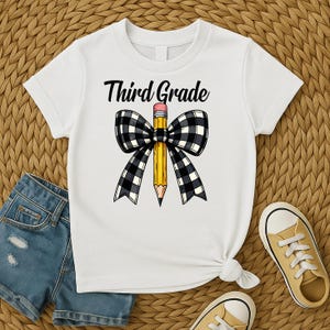 Puede incluir: Camiseta blanca con la frase "Third Grade" sobre un lápiz amarillo con goma rosa, envuelta en un lazo a cuadros blanco y negro. La camiseta se combina con pantalones cortos de mezclilla y zapatillas amarillas.