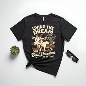 May include: Black t-shirt with a graphic featuring a cartoon goat and raccoon on a wooden fence. The text reads "LIVING THE DREAM" above and "ONE BAD DECISION AT A TIME" below the image. A small succulent and sunglasses are also visible.