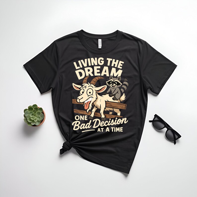 May include: Black t-shirt with a graphic featuring a cartoon goat and raccoon on a wooden fence. The text reads "LIVING THE DREAM" above and "ONE BAD DECISION AT A TIME" below the image. A small succulent and sunglasses are also visible.