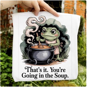 May include: White tote bag featuring a cartoon frog chef stirring a pot of soup. The frog wears a pink apron and the text reads "That's it. You're Going in the Soup."