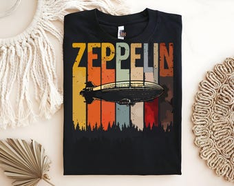 Può includere: T-shirt nera con la scritta "ZEPPELIN" in giallo, bianco e arancione, sopra un'illustrazione vintage di un dirigibile. Il design include uno sfondo a strisce colorate e una silhouette di foresta.