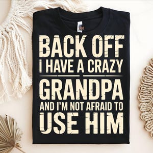 May include: Black t-shirt with the text "BACK OFF I HAVE A CRAZY GRANDPA AND I'M NOT AFRAID TO USE HIM" in a bold, off-white font. The shirt is displayed on a neutral surface with decorative elements.