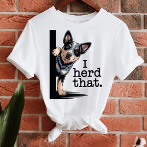 May include: White t-shirt featuring a cartoon Blue Heeler dog peeking from behind a black vertical line. The text "I herd that." is printed in black. The shirt is hanging on a wooden hanger against a brick wall.