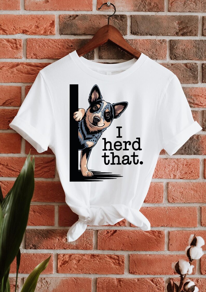 May include: White t-shirt featuring a cartoon Blue Heeler dog peeking from behind a black vertical line. The text "I herd that." is printed in black. The shirt is hanging on a wooden hanger against a brick wall.