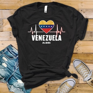 Może przedstawiać: Czarna koszulka z sercem w kolorach wenezuelskiej flagi i grafiką linii życia. Słowo "VENEZUELA" jest wydrukowane na biało, wraz z hasztagiem "#LIBRE".