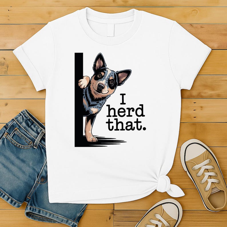May include: White t-shirt featuring a blue heeler dog peeking from behind a black vertical line. The text "I herd that." is printed in black. The dog has a black and blue pattern. The shirt is on a wooden surface.