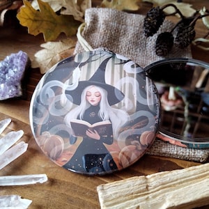 Könnte beinhalten: Ein runder, kompakter Spiegel mit einer Hexe, die in einer Waldkulisse ein Buch liest. Der Spiegel hat einen silbernen Rahmen und einen schwarzen Hintergrund. Die Hexe trägt ein schwarzes Kleid und einen spitzen Hut. Das Buch hat einen goldenen Einband und der Text "Book of Shadows" ist sichtbar.