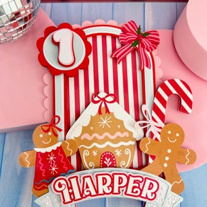 May include: A colourful cake topper featuring a gingerbread house, two gingerbread people, and a red and white striped backdrop. The number "1" is displayed, along with the name "HARPER" on a banner. A candy cane and a pink bow complete the design.