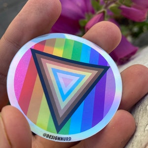 May include: A round, iridescent sticker with a rainbow-colored stripe pattern and a black triangle with concentric circles of different shades of brown in the center. The sticker has the text "@DESIGNNURD" at the bottom.
