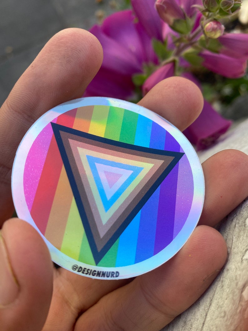May include: A round, iridescent sticker with a rainbow-colored stripe pattern and a black triangle with concentric circles of different shades of brown in the center. The sticker has the text "@DESIGNNURD" at the bottom.