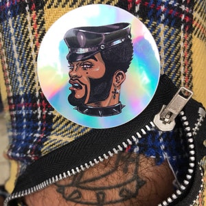 May include: A holographic sticker featuring a cartoon illustration of a person with a black leather cap, a black beard, and a silver nose ring. The person is wearing a black leather collar with silver studs.