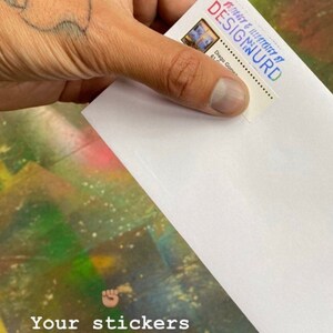May include: A white envelope with a sticker on the front that says "DESIGNED & ILLUSTRATED BY DESIGWURD". The text "Your stickers will come in an envelope like this" is written below the envelope. A rainbow emoji is below the text.