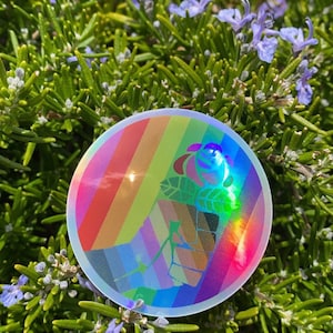 Puede incluir: Una pegatina holográfica arcoíris con un puño en alto sosteniendo una rosa. La pegatina es redonda y tiene un borde blanco.