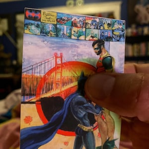 May include: A Batman and Robin comic art print. The print features Batman and Robin in a scene with the Golden Gate Bridge in the background. The top of the print has a Batman logo and comic panels. The colors are vibrant.