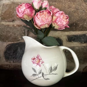 May include: A white ceramic jug with a pink rose design, holding a bouquet of pink and cream artificial peonies. The jug has a round shape with a handle and a spout. The background is a stone wall.