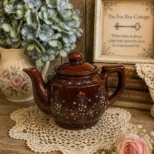 May include: A glossy brown ceramic teapot featuring a hand-painted floral pattern with small orange and blue flowers, a matching lid, a curved spout, and a sturdy handle. The teapot is decorated with delicate white and blue botanical details.