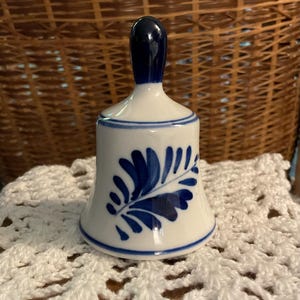 May include: A white ceramic bell with a blue handle and leaf design. The bell has a blue rim and a white body with a blue floral pattern. It is sitting on a white crocheted doily.
