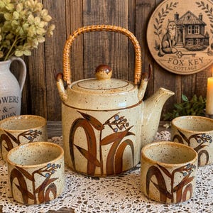 May include: A ceramic tea set featuring a speckled beige teapot with a woven rattan handle and three matching cups. Each piece is decorated with hand-painted brown bamboo stalks and small blue berries. The teapot includes a lid with a knob.
