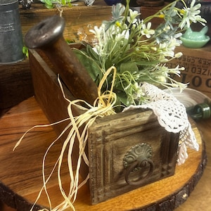 May include: A rustic wooden box with a metal handle and a decorative floral arrangement. The box is filled with white and green flowers, and accented with a lace doily and raffia. The box sits on a wooden slice.