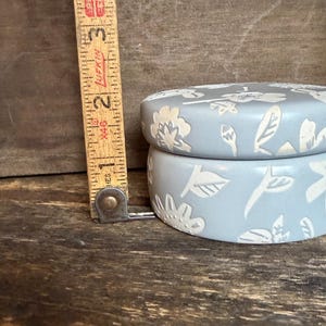 Może przedstawiać: Jasnoniebieskie ceramiczne pudełko na drobiazgi z kwiatowym wzorem. Pudełko ma średnicę około 7,6 cm i wysokość 5 cm. Pokrywka i boki zdobione są białymi, wytłaczanymi kwiatami i liśćmi. Pudełko stoi na drewnianej powierzchni.
