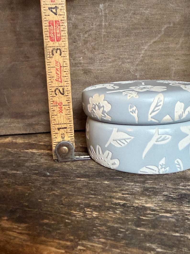 Może przedstawiać: Jasnoniebieskie ceramiczne pudełko na drobiazgi z kwiatowym wzorem. Pudełko ma średnicę około 7,6 cm i wysokość 5 cm. Pokrywka i boki zdobione są białymi, wytłaczanymi kwiatami i liśćmi. Pudełko stoi na drewnianej powierzchni.