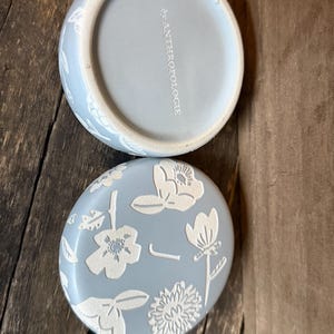 Może przedstawiać: Jasnoniebieskie ceramiczne pudełko na drobiazgi z pokrywką z białymi kwiatowymi wzorami. Pokrywka jest okrągła i ma zakrzywioną krawędź. Sp&oacute;d pudełka jest jasnoniebieski z białym napisem "by ANTHROPOLOGIE".