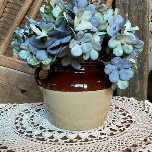 May include: A brown and beige ceramic jug with a handle, filled with blue and green artificial flowers. The jug sits on a white crocheted doily. The background includes wooden elements.