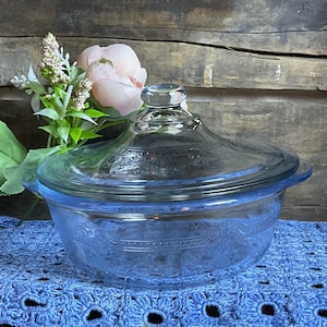 May include: A vintage, light blue glass casserole dish with a clear glass lid. The dish has a rounded shape with handles and decorative embossed patterns. The lid has a knob handle. The dish is sitting on a blue crocheted surface.