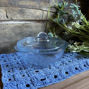 Peut inclure: Un plat à casserole en verre bleu clair avec un couvercle assorti, posé sur un napperon bleu crocheté. Le plat présente un motif décoratif et une poignée en boucle sur le couvercle. Des fleurs décoratives sont en arrière-plan.