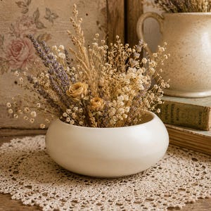May include: A white ceramic bowl filled with dried flowers, including lavender and wheat, sits on a crocheted doily. A beige pitcher with dried flowers and a vintage book are in the background. The arrangement is on a wooden surface.