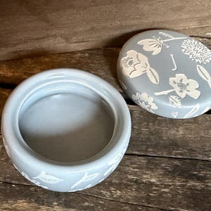 Może przedstawiać: Jasnoniebieskie ceramiczne pudełko na drobiazgi z pasującą pokrywką. Pokrywka jest ozdobiona białymi wzorami kwiatowymi i owadami oraz literą "J". Pudełko ma okrągły kształt z pasującym kwiatowym wzorem na podstawie. Pudełko jest otwarte, odsłaniając wnętrze.