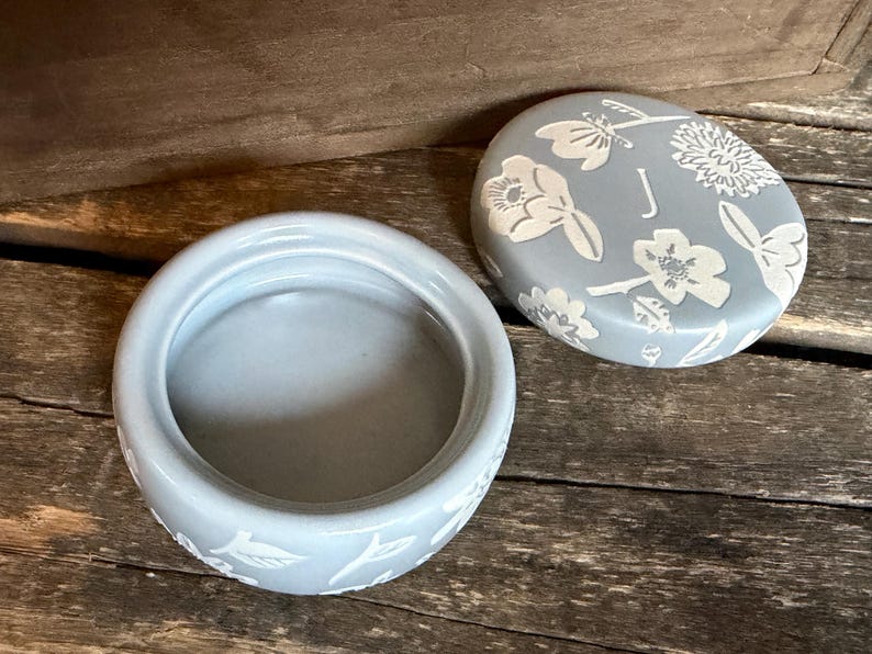 Może przedstawiać: Jasnoniebieskie ceramiczne pudełko na drobiazgi z pasującą pokrywką. Pokrywka jest ozdobiona białymi wzorami kwiatowymi i owadami oraz literą "J". Pudełko ma okrągły kształt z pasującym kwiatowym wzorem na podstawie. Pudełko jest otwarte, odsłaniając wnętrze.