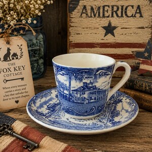 May include: A white ceramic teacup and saucer set featuring a blue transferware pattern depicting Mount Vernon. The cup and saucer display detailed architectural illustrations and text. A metal key, a tag labeled The Fox Key Cottage, and patriotic decor are present.