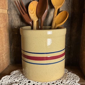 May include: A tan ceramic utensil holder with blue and red stripes, filled with wooden spoons and a fork. The holder sits on a white lace doily, with a rustic wooden background.