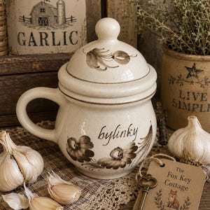 May include: A cream-colored ceramic jar with a lid and handle, featuring brown floral patterns and the word "bylinky" printed on the front. A brown tag labeled "The Fox Key Cottage" with a fox illustration and a metal key are attached to the side.