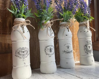 May include: Four white painted glass bottles with distressed paint and black and white vintage labels. Each bottle is filled with dried lavender and tied with twine. The labels feature bees, butterflies, and floral designs.