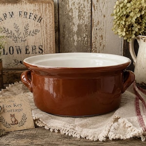 May include: A round, glossy brown ceramic casserole dish with two side handles and a smooth white interior. A small rectangular tag is attached to the dish, featuring a drawing of a fox and the text, The Fox Key Cottage, along with a metal key.